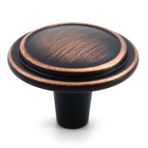 May include: A round cabinet knob featuring a dark oil-rubbed bronze finish with copper-toned highlights along the edges. The knob has a smooth, slightly domed top surface and a tapered cylindrical base, providing a functional hardware piece.