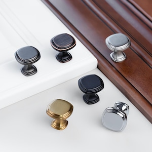 May include: Six square cabinet knobs in different finishes: oil rubbed bronze, brushed nickel, black, polished nickel, and gold. The knobs are displayed on a white and wood surface.