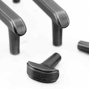 May include: A set of dark gray metal cabinet pulls and a single T-shaped knob. Each piece features a brushed finish with fine horizontal ribbed detailing along the top edges and a smooth, cylindrical base for mounting to furniture or cabinetry.
