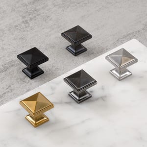 May include: Five square cabinet knobs in various metallic finishes: black, bronze, silver, gold, and a darker bronze. Each knob has a pyramidal top and a square base, suitable for drawers or cabinet doors.