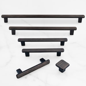 May include: A collection of dark bronze cabinet hardware featuring four linear bar pulls of varying lengths, one angled bar pull, and one square cabinet knob. Each piece has a brushed metallic finish with rounded edges and two mounting posts.