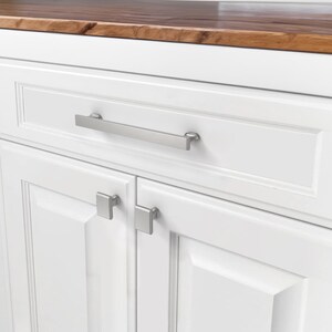May include: Close-up of a white cabinet with a wooden countertop. The cabinet has a long, brushed nickel handle on the drawer and square knobs on the doors. The wood countertop has a rich, dark brown color.