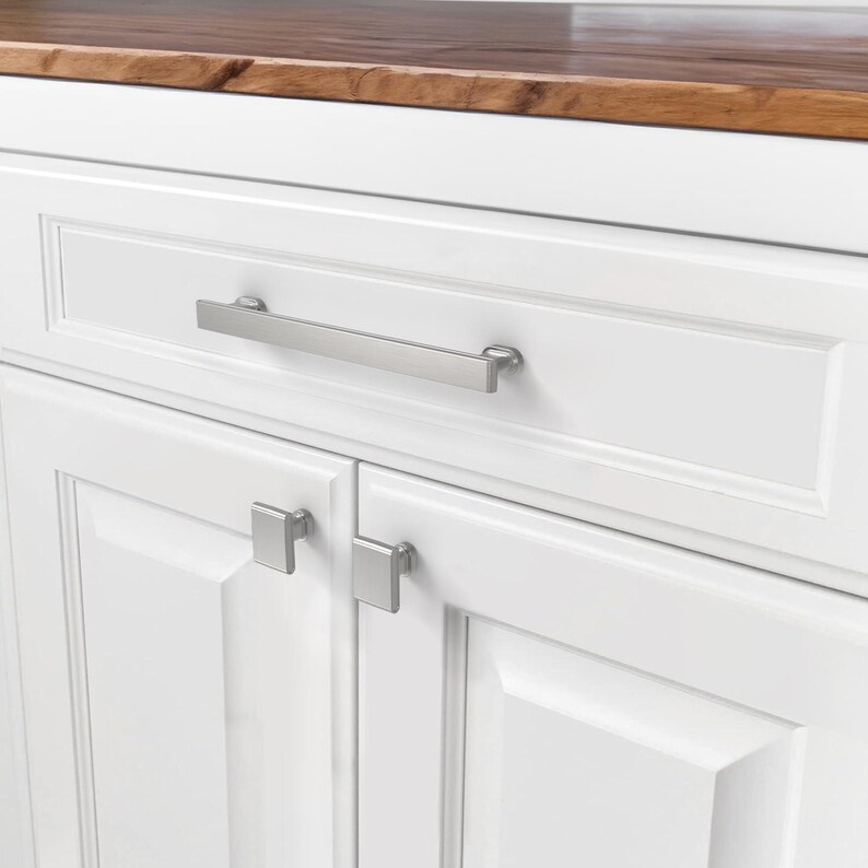 May include: Close-up of a white cabinet with a wooden countertop. The cabinet has a long, brushed nickel handle on the drawer and square knobs on the doors. The wood countertop has a rich, dark brown color.