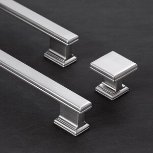 May include: Two long, rectangular cabinet pulls and one square cabinet knob made of brushed silver metal. Each piece features a stepped base design with clean, straight edges and a smooth, metallic finish that reflects light across the surface.
