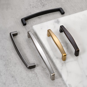 May include: An assortment of cabinet pulls in various finishes, including black, silver, gold, and bronze. The pulls are displayed on a white marble surface and a gray background. The hardware features a variety of shapes and styles.