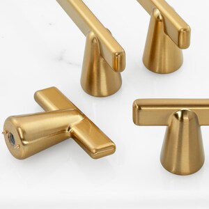 May include: A set of four T-shaped cabinet knobs with a brushed gold finish. Each knob features a conical base that connects to a horizontal bar handle. The metal surface has a smooth, matte texture, and the base shows a threaded mounting hole.