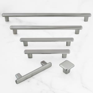May include: A collection of brushed nickel cabinet hardware, including several sizes of bar pulls and a square knob. The hardware is displayed on a white marble surface, showcasing the variety of options available for kitchen or bathroom cabinetry.