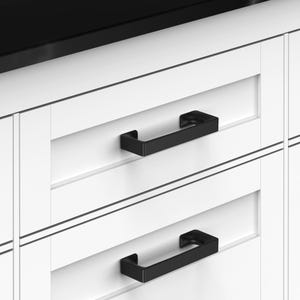 May include: Two matte black rectangular cabinet pulls are mounted horizontally on white shaker-style drawers. The hardware features a clean, geometric design with sharp edges and a solid construction, contrasting against the bright white cabinetry.