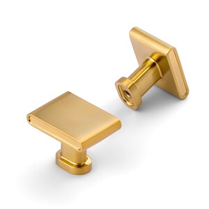 May include: Two gold-colored cabinet knobs with a rectangular face and rounded edges. The knobs have a brushed metal finish and a cylindrical base. The hardware is designed for easy installation and adds a touch of elegance to any furniture piece.