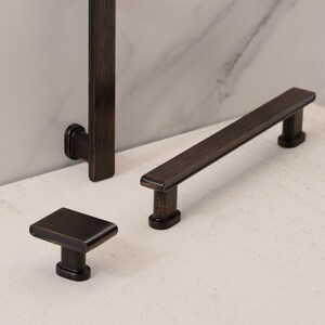 May include: A collection of bronze-toned cabinet hardware, including a square knob, a long bar pull, and a vertical handle. The hardware has a sleek, modern design with a dark, metallic finish, suitable for kitchen or bathroom cabinetry.
