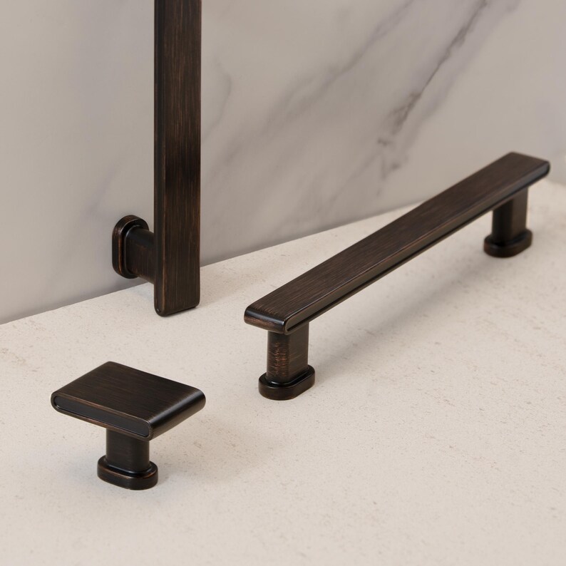 May include: A collection of bronze-toned cabinet hardware, including a square knob, a long bar pull, and a vertical handle. The hardware has a sleek, modern design with a dark, metallic finish, suitable for kitchen or bathroom cabinetry.