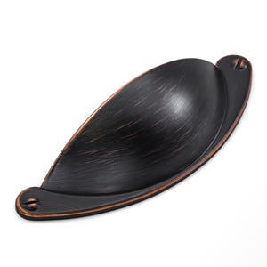 May include: A dark oil-rubbed bronze cup pull with a brushed finish and copper-toned edges. The hardware features a curved, shell-like shape with two integrated mounting points at the ends, each containing a single visible screw head.