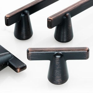 May include: A set of T-shaped cabinet pulls featuring a dark bronze finish with copper-toned highlights on the edges. Each pull consists of a horizontal bar attached to a conical base, showcasing a brushed metal texture throughout the surface.