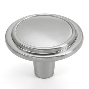 May include: A circular cabinet knob with a brushed silver finish. The knob features a smooth, slightly domed centre disc surrounded by a raised, rounded outer ring, connected to a tapered cylindrical base for mounting to a drawer or door.