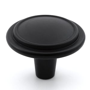 May include: A matte black cabinet knob featuring a circular, slightly domed top with a raised outer rim. The knob has a tapered, cylindrical base that extends downwards, providing a solid, uniform appearance for furniture or cabinetry hardware.