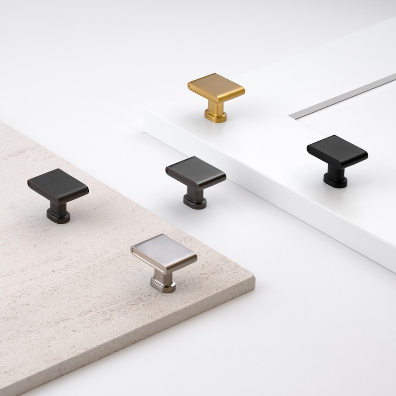 May include: A collection of rectangular cabinet knobs in various metallic finishes: gold, black, dark gray, silver, and a lighter gray. The knobs are displayed on a white and beige surface, showcasing their design and color variations.