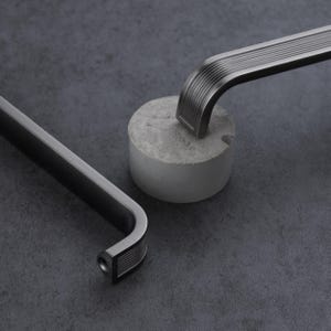 May include: Two dark gray metal cabinet handles with a ribbed texture on the curved ends. Each handle features a long, smooth central bar and a mounting hole at the base. The hardware has a matte finish and a sleek, elongated rectangular shape.