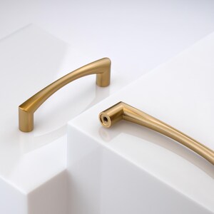 May include: Two curved cabinet pulls with a brushed gold finish. Each handle features a smooth, arched design with cylindrical mounting points at both ends. The end of one handle is visible, showing a circular opening with a central screw hole.