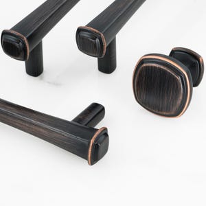 May include: A collection of dark bronze cabinet hardware featuring three elongated bar pulls and one square knob. Each piece has a brushed finish with copper-toned edges, a rounded square profile, and a central mounting post for attachment.