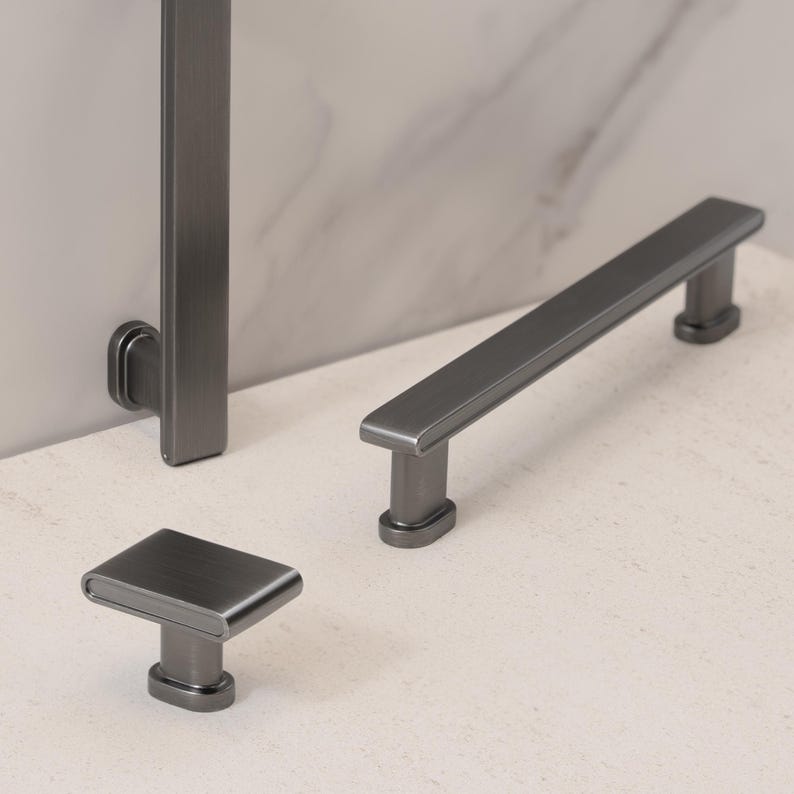 May include: A set of dark gray cabinet hardware, including a rectangular knob, a long bar pull, and a vertical handle. The hardware has a brushed metal finish and a modern design. The pieces are displayed on a light-colored surface.