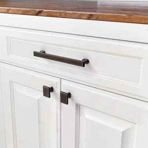 May include: Close-up of white cabinetry with dark bronze hardware. The drawer features a long, rectangular pull, while the cabinet doors below have square knobs. The countertop is a warm, natural wood tone.