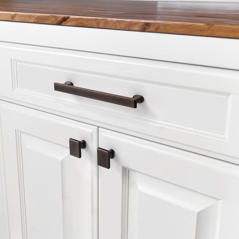 May include: Close-up of white cabinetry with dark bronze hardware. The drawer features a long, rectangular pull, while the cabinet doors below have square knobs. The countertop is a warm, natural wood tone.