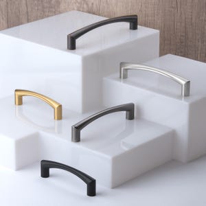 May include: An assortment of cabinet handles in various metallic finishes, including gold, silver, and black. The handles are arched and mounted on white platforms, showcasing their design and texture. The handles are designed for furniture.