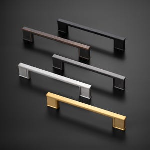 May include: A selection of cabinet pulls in various metallic finishes, including black, bronze, silver, and gold. Each pull features a rectangular bar design with square end caps, set against a dark background.