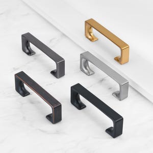 Może przedstawiać: Pięć gałek do szafek w różnych wykończeniach: czarny, brązowy, srebrny i złoty. Gałki mają ten sam kształt, z prostokątną podstawą i zakrzywionym wierzchołkiem.