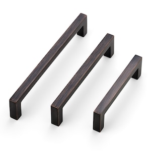 May include: Three bronze-colored cabinet pulls in varying sizes. Each pull has a rectangular bar with a square, right-angled base. The hardware is displayed against a white background, showcasing the details of the metal finish.