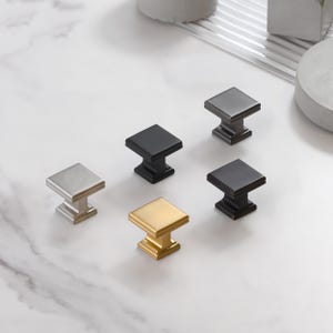 May include: An assortment of square cabinet knobs in various metallic finishes, including silver, black, gold, and bronze. The knobs are arranged on a white marble surface, showcasing their different colors and designs.