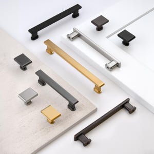 May include: A collection of cabinet hardware, including various pull handles and square knobs. Finishes include matte black, brushed nickel, gold, and bronze. The pulls are rectangular, and the knobs are square.