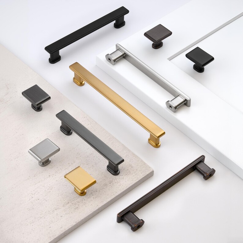 May include: A collection of cabinet hardware, including various pull handles and square knobs. Finishes include matte black, brushed nickel, gold, and bronze. The pulls are rectangular, and the knobs are square.