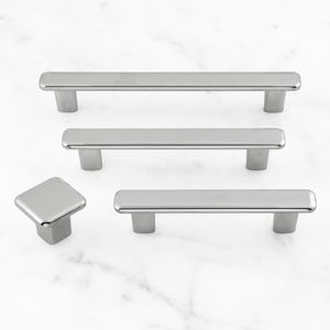 May include: A collection of four silver-toned metal cabinet hardware pieces, including three rectangular bar pulls of varying lengths and one square-shaped knob, all featuring a smooth, polished finish and rounded edges for a clean look.