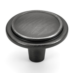 Puede incluir: Un pomo redondo para muebles con un acabado metálico oscuro y cepillado. El pomo presenta una superficie superior plana y circular con una textura cepillada sutil, rodeada por un borde exterior liso y elevado, y una base cilíndrica cónica.
