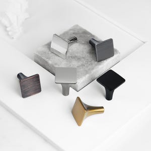 May include: Five different square cabinet knobs in various finishes, including brushed nickel, brass, copper, and black. The knobs are displayed on a light gray stone surface.