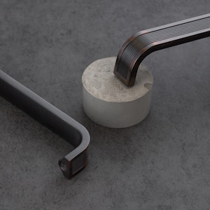 May include: Two metal cabinet pulls with a dark, brushed finish and a ribbed, textured design. One pull is matte black, while the other features a warm, bronze-toned metallic sheen. Both handles have a curved, elongated shape with visible screw holes.
