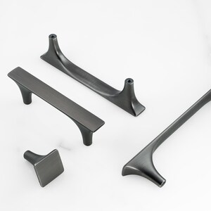 May include: A collection of dark gray metal cabinet hardware including three elongated drawer pulls with curved supports and one square-shaped cabinet knob. Each piece features a brushed finish and visible screw holes for mounting purposes.
