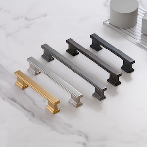 May include: An assortment of cabinet pulls in various metallic finishes, including gold, silver, and dark gray. The pulls have a rectangular bar design with square end supports. The hardware is displayed on a white marble surface.