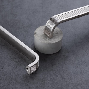 May include: Two silver-toned metal cabinet pulls with a brushed finish. Each handle features a curved end with a series of parallel horizontal grooves. The design includes a flat, elongated bar section and a mounting point with a visible screw hole.