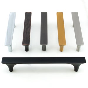 May include: Six different finishes of a modern cabinet pull. The pulls are rectangular with rounded edges and a slight curve at the bottom. The finishes are polished nickel, oil rubbed bronze, brushed nickel, gold, satin nickel, and black.