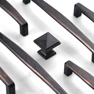 May include: A collection of dark bronze cabinet hardware featuring long, straight drawer pulls with rectangular mounting posts and a single square cabinet knob with a pyramid-shaped top, all displaying a brushed metallic finish.