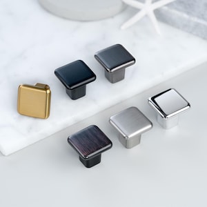 Pode incluir: Uma coleção de puxadores quadrados para armários em vários acabamentos metálicos, incluindo dourado, preto, bronze e prata. Os puxadores estão dispostos sobre uma superfície branca, mostrando suas diferentes cores e texturas. Ideais para armários.