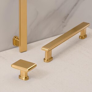 May include: A collection of gold-colored cabinet hardware, including a rectangular knob, a long bar pull, and a vertical pull. The hardware is displayed on a light-colored surface with a marble-patterned background.