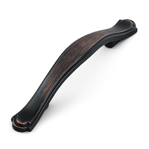 May include: A curved cabinet pull handle featuring a dark oil-rubbed bronze finish with copper-toned highlights along the edges and center. The hardware has a smooth, arched design with decorative flared ends and two mounting points on the underside.