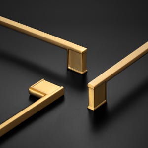 May include: Three gold-colored metal cabinet pulls with a linear, rectangular design. Each handle features a long, slender bar attached to a solid, block-shaped mounting base at one end, showcasing a smooth, brushed metallic finish throughout.