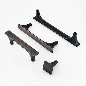May include: An assortment of cabinet hardware, including handles and a knob, in a mix of bronze and black finishes. The handles have a sleek, modern design with a curved shape and a textured surface. The hardware is designed for furniture.