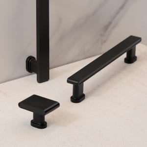 May include: A collection of matte black cabinet hardware, including a square knob, a rectangular pull, and a vertical handle. The hardware is displayed on a light-colored surface, showcasing its sleek, modern design.
