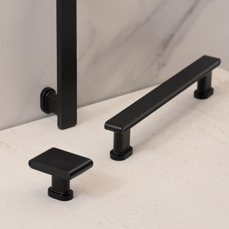 May include: A collection of matte black cabinet hardware, including a square knob, a rectangular pull, and a vertical handle. The hardware is displayed on a light-colored surface, showcasing its sleek, modern design.