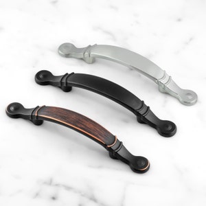 Op de afbeelding: Drie kastgrepen in verschillende afwerkingen: olie gebronsd, zwart en nikkel. Elke greep heeft een gebogen vorm met een afgerond uiteinde.